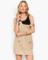 A woman modeling a beige corduroy pinafore mini dress over a black ribbed tank top, set against a plain white studio background.