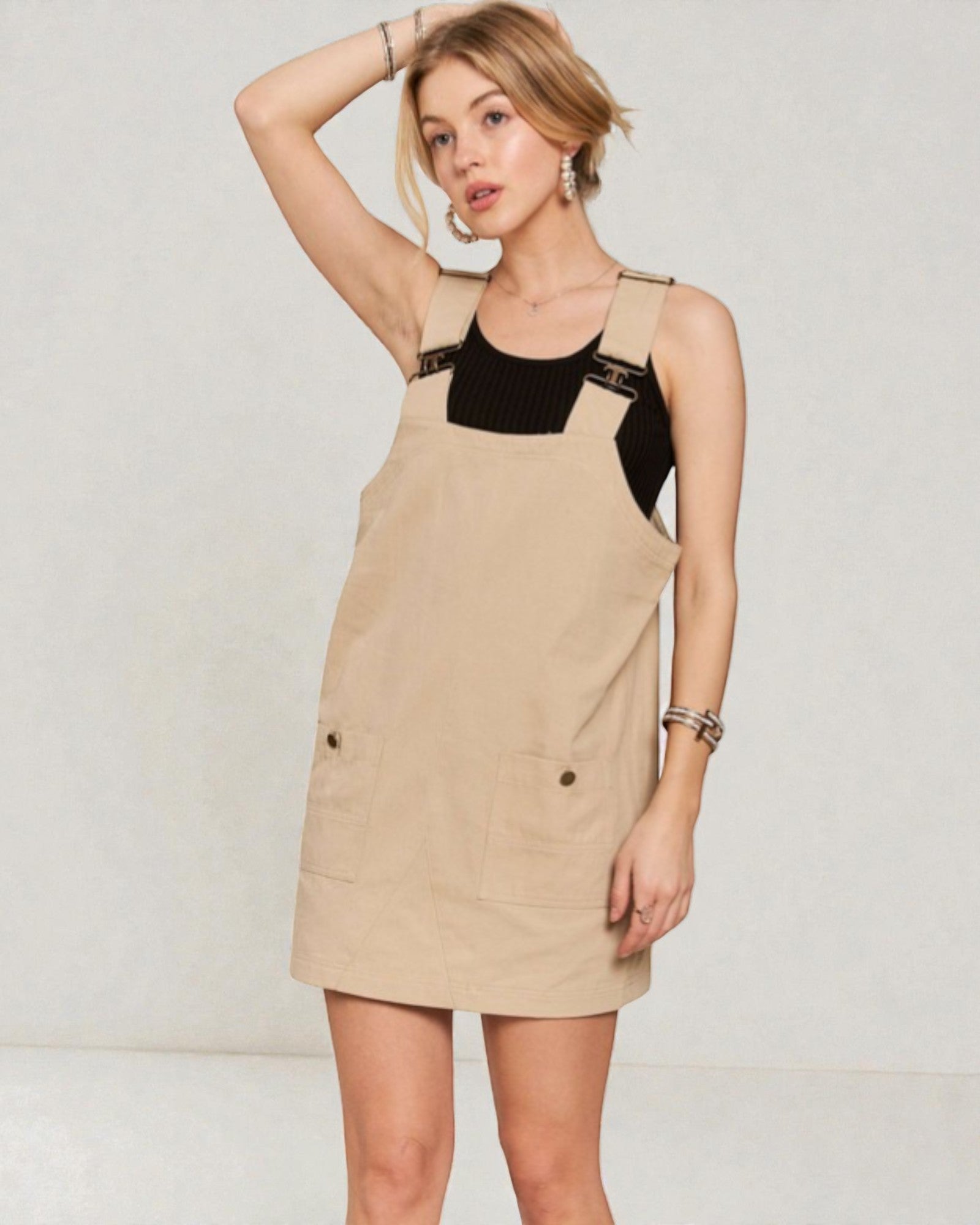 Woman wearing a beige pinafore dress with black top against a plain background