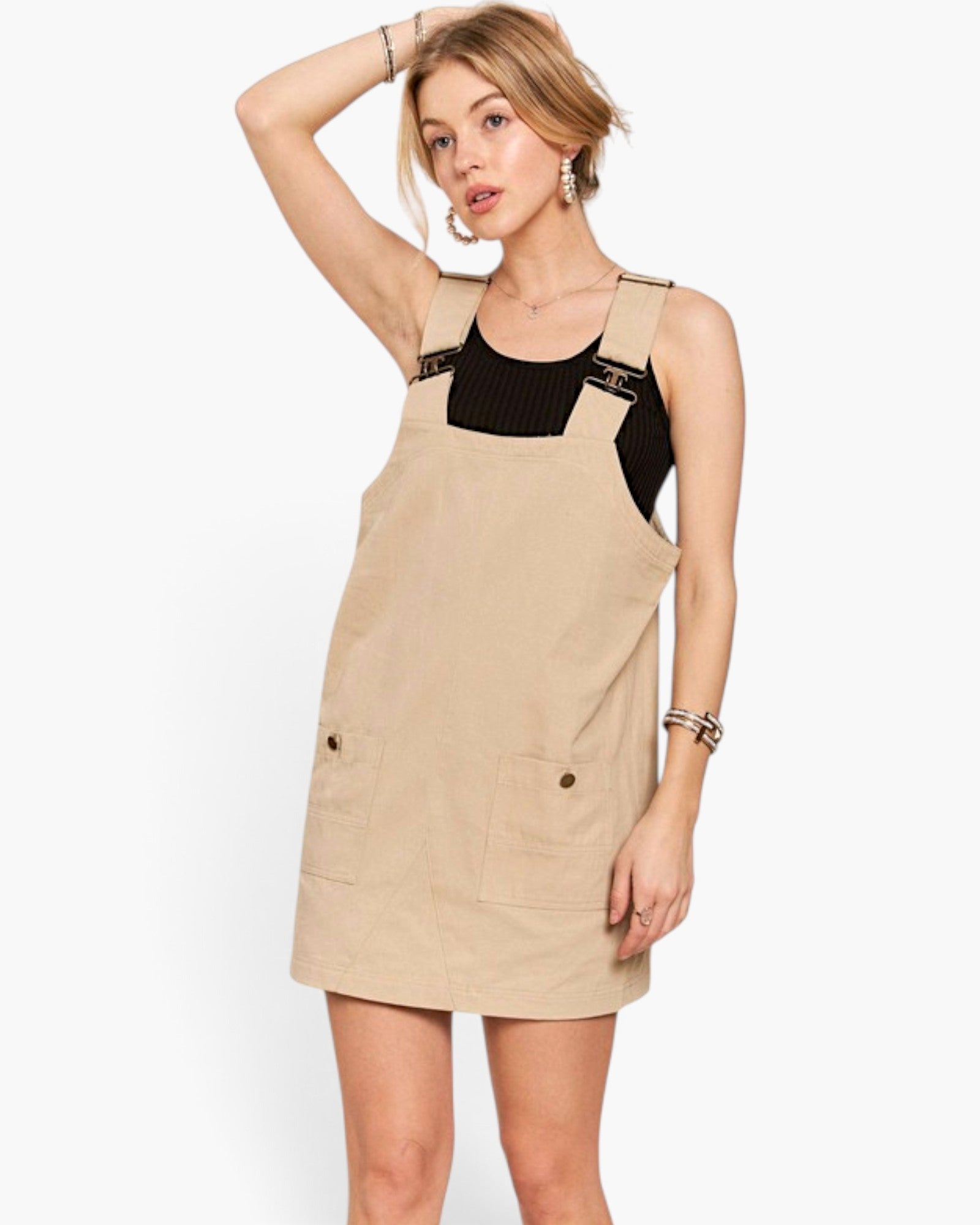 A woman modeling a beige corduroy pinafore dress over a black ribbed tank top, photographed against a clean white studio background.