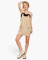 A woman modeling a beige pinafore-style mini dress with black shoulder straps and front patch pockets, paired with beige lace-up ankle boots, on a light gray studio background.