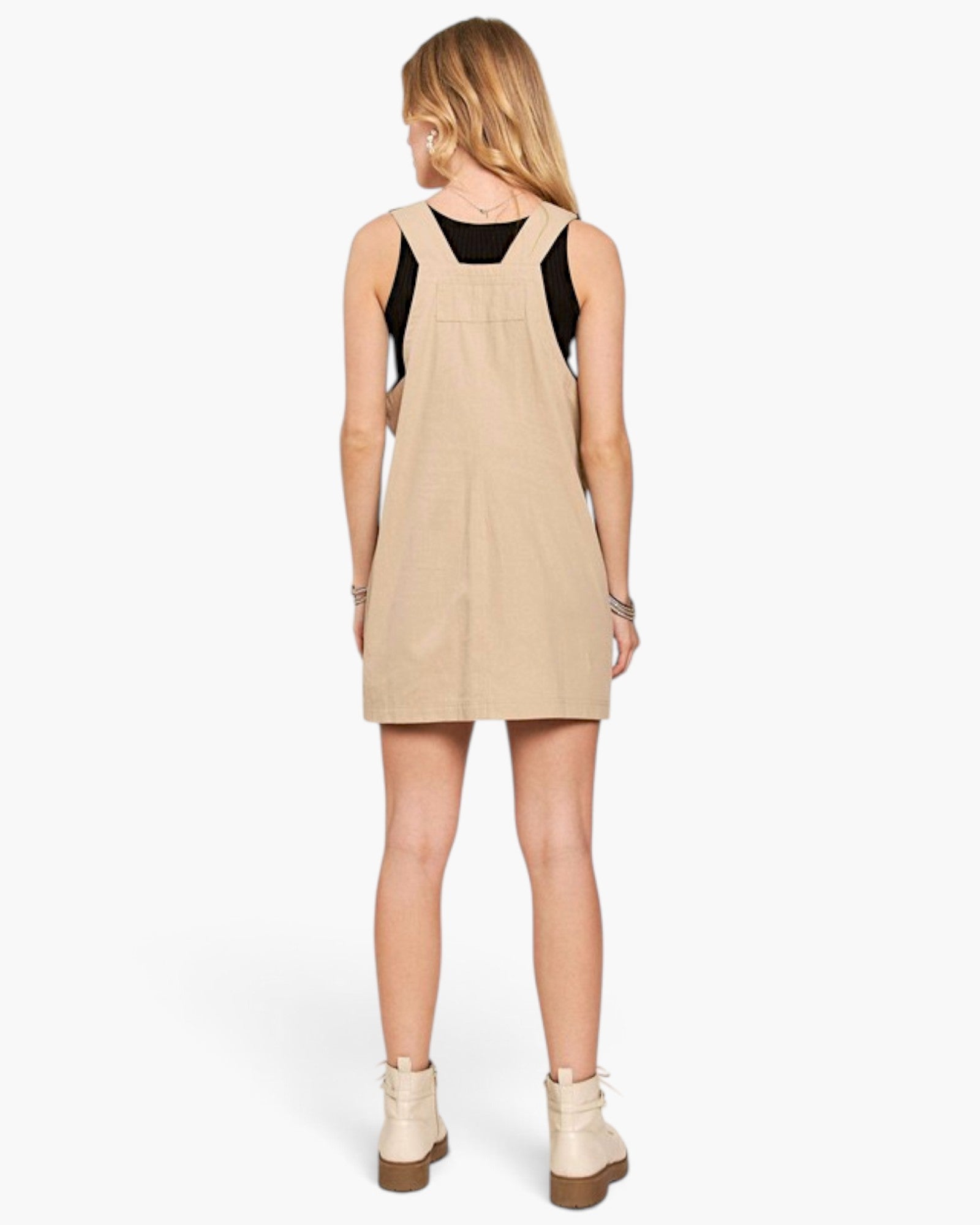 A woman shown from the back wearing a beige pinafore-style mini dress over a black sleeveless top, paired with cream ankle boots, against a light gray studio background.