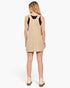 A woman shown from the back wearing a beige pinafore-style mini dress over a black sleeveless top, paired with cream ankle boots, against a light gray studio background.
