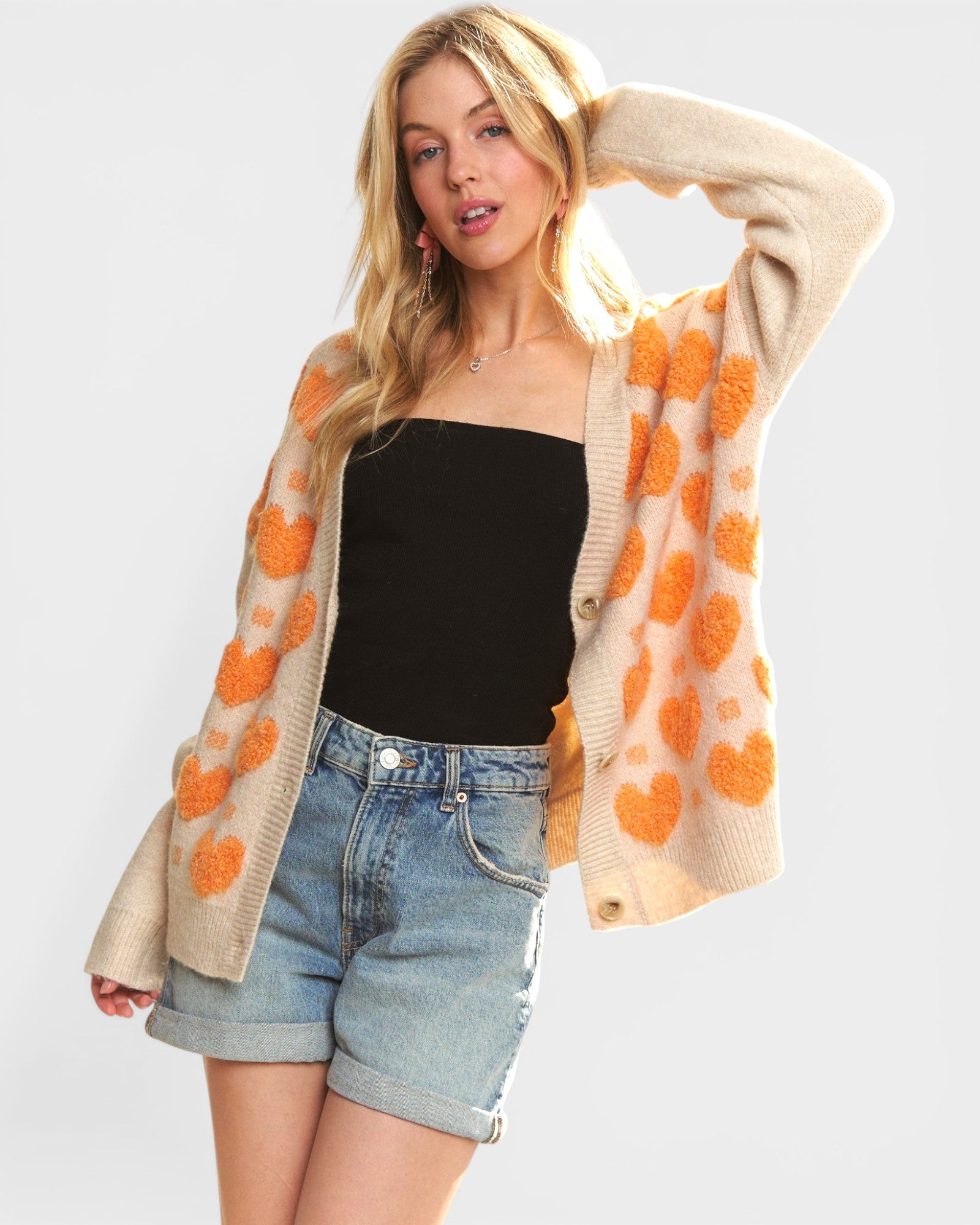 Woman wearing a beige cardigan with orange heart patterns, black top, and denim shorts on a white background