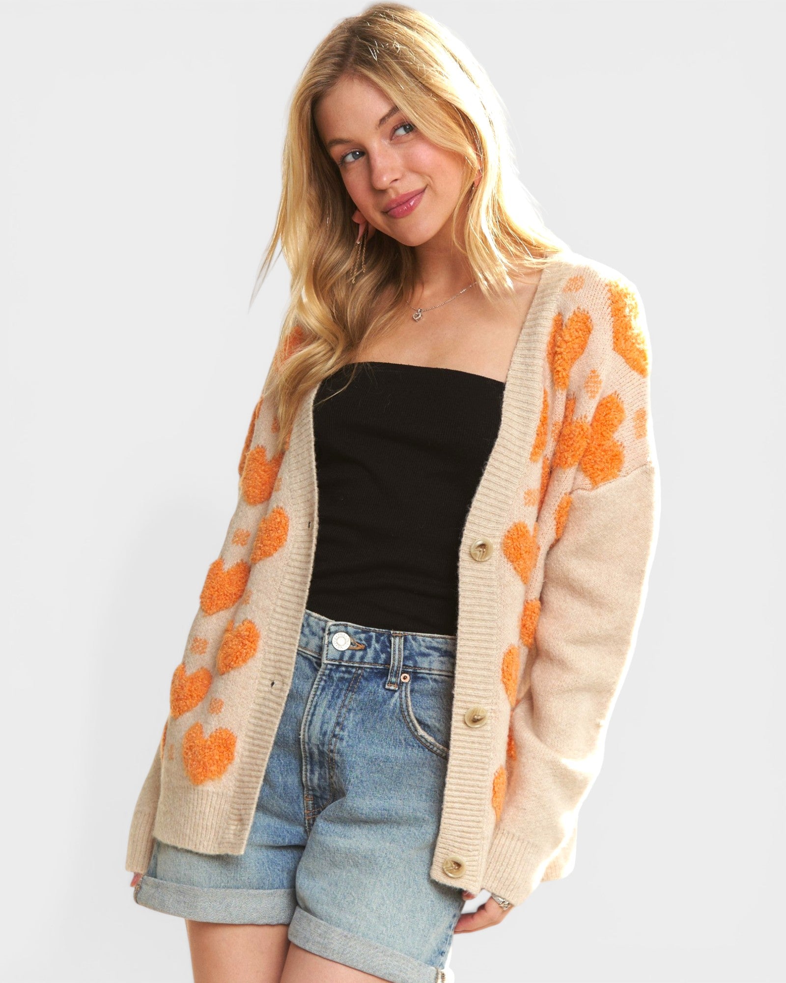 Woman wearing a beige cardigan with orange floral patterns, black top, and denim shorts on a white background