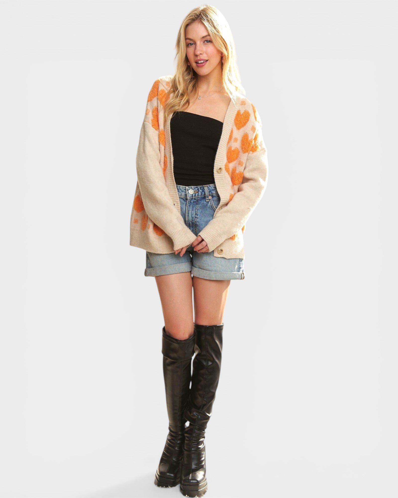 Woman wearing a patterned cardigan, black top, denim shorts, and knee-high boots on a white background