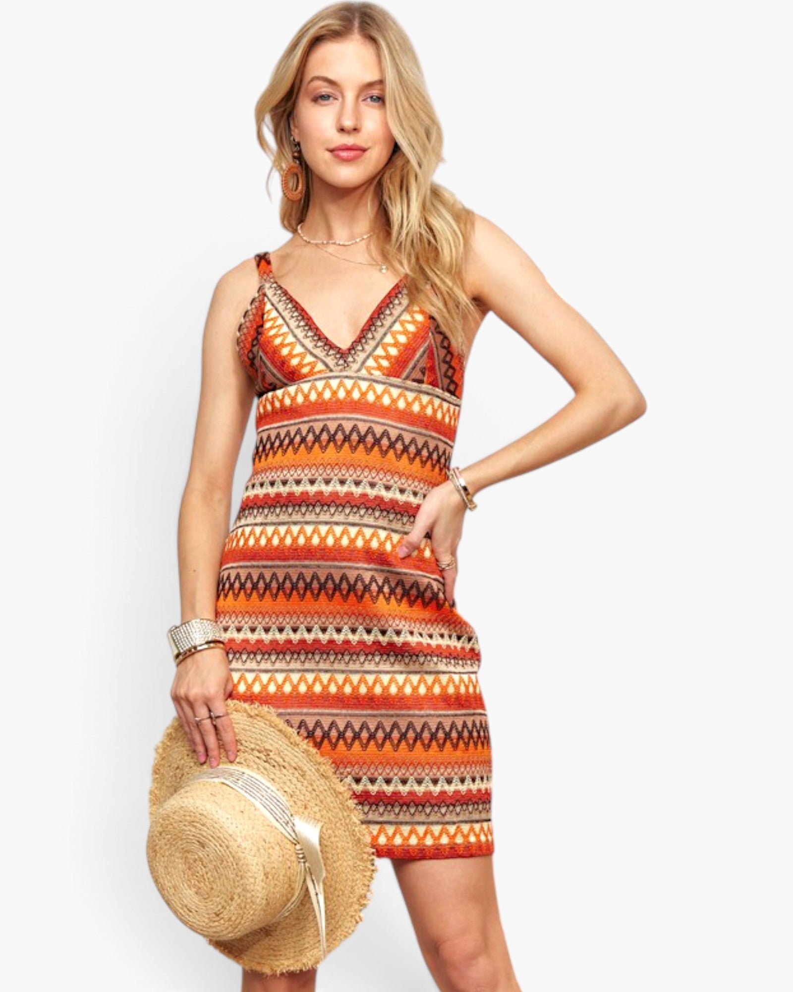 A woman modeling a sleeveless V-neck mini dress with a colorful geometric tribal pattern in orange, red, black, and beige. She is holding a straw hat against a plain white studio background.