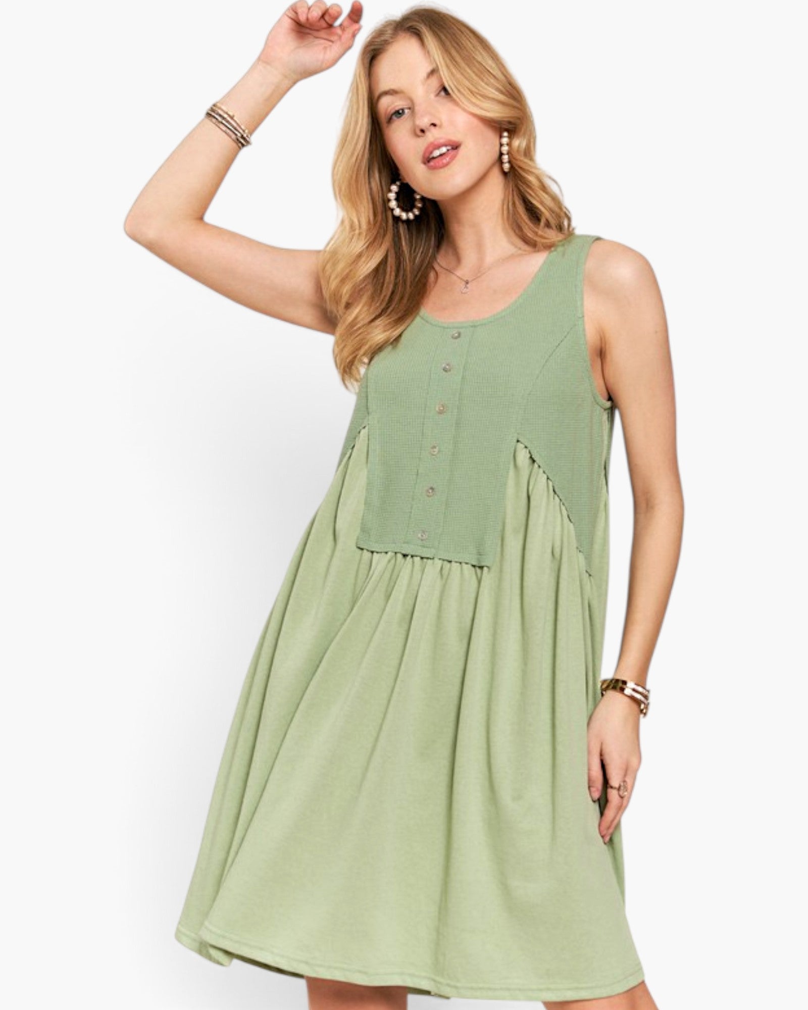 A woman modeling a sleeveless light sage green dress with a buttoned bodice and gathered skirt, posed against a clean white studio background.