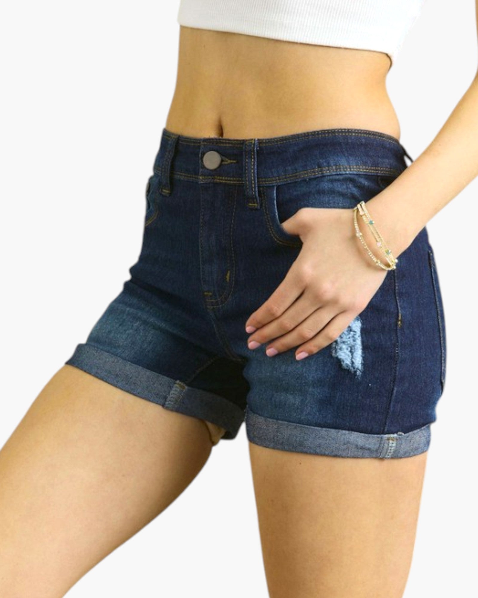 A close-up of a model wearing dark blue denim shorts with a distressed rip on the left leg and rolled hems, paired with a white crop top against a plain white background.