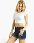 A woman modeling a white short-sleeve crop top and dark blue denim shorts against a plain white background.