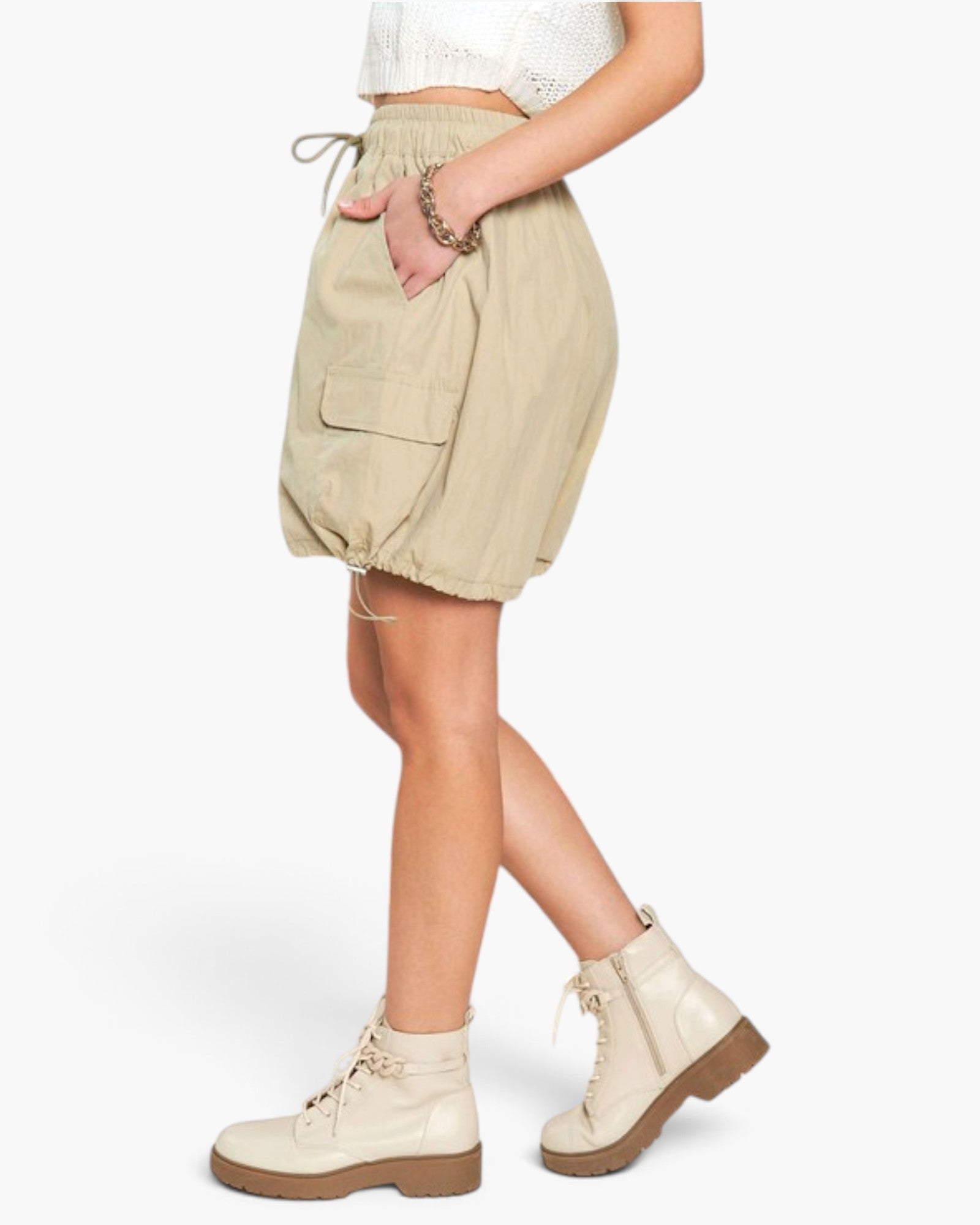 A model wearing beige cargo-style biker shorts with a drawstring waist, side pockets, and a gathered elastic hem, styled with cream combat boots against a plain white background.