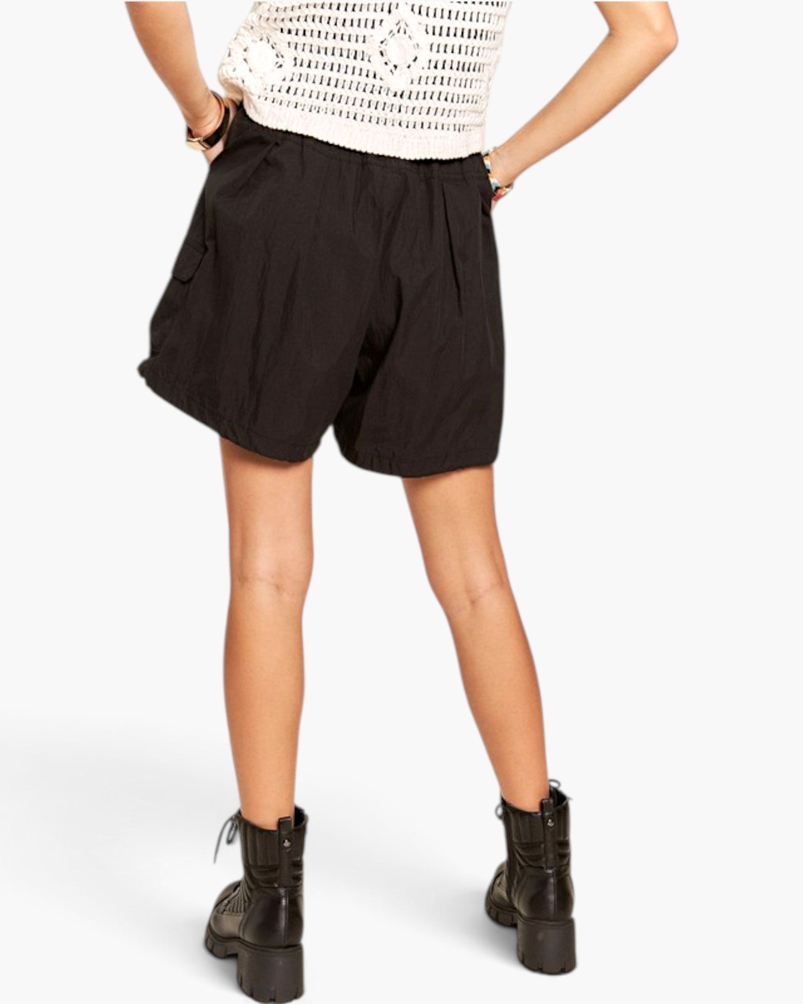 A model shown from the back wearing black cargo shorts, a white crochet knit top, and black lace-up ankle boots against a light gray studio background.