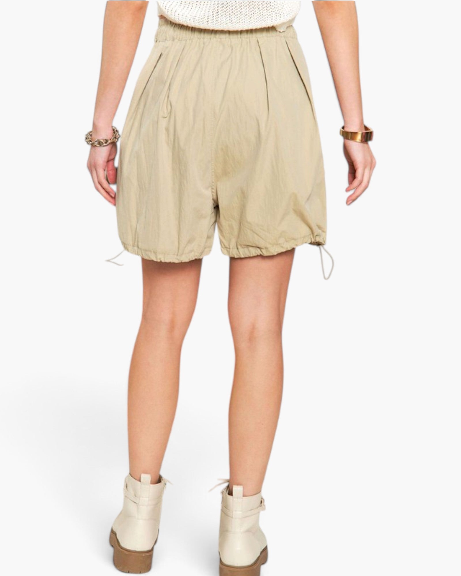 A model from the back wearing beige high-waisted drawstring shorts, a white knit top, and off-white platform ankle boots against a light gray studio background.
