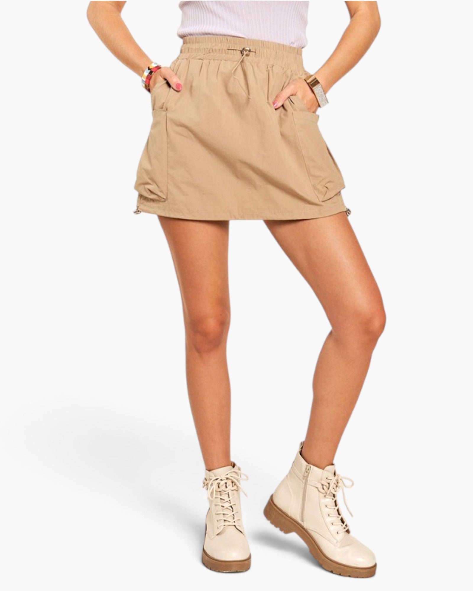 A model wearing a beige utility-style mini skirt with side pockets, paired with cream lace-up boots, shown against a plain light gray studio background.