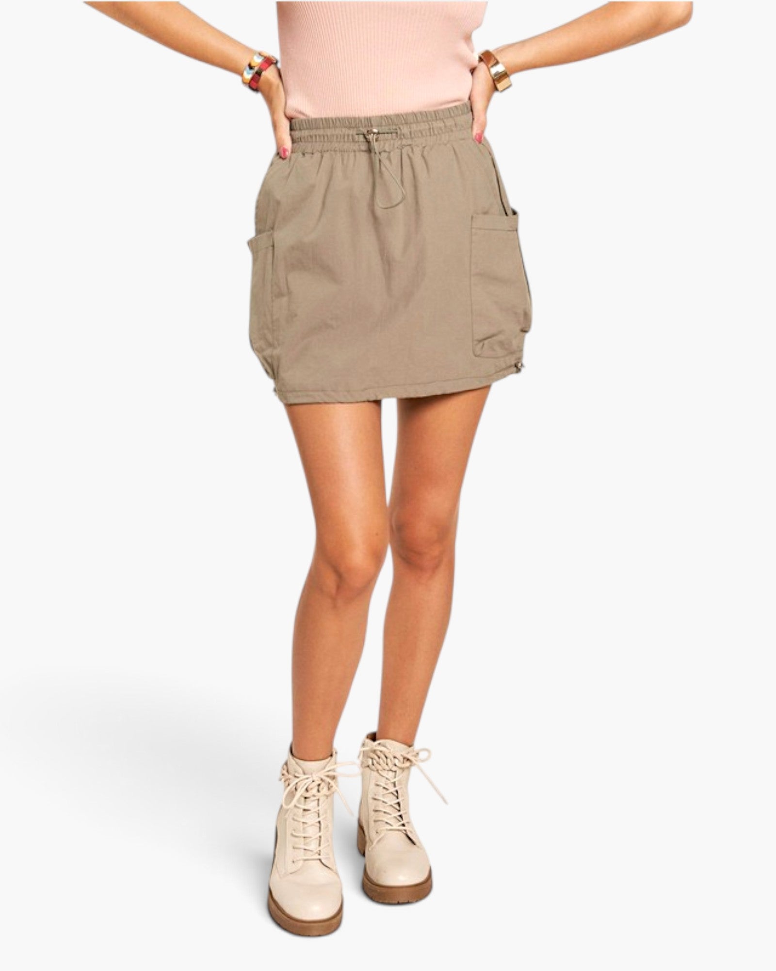 A model wearing a light brown utility mini skirt with side pockets, paired with a pink top and beige lace-up boots, shown from the waist down on a white studio background.