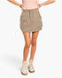 A model wearing a light brown utility mini skirt with side pockets, paired with a pink top and beige lace-up boots, shown from the waist down on a white studio background.