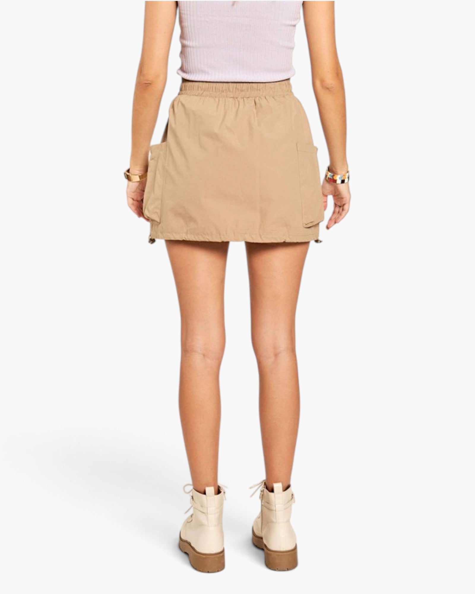 A model from the back wearing a beige cargo mini skirt, a light pink ribbed top, and beige ankle boots, set against a plain white studio background.