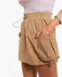 A close-up of a person wearing tan drawstring shorts with side pockets, paired with a light pink ribbed top, against a plain white background.