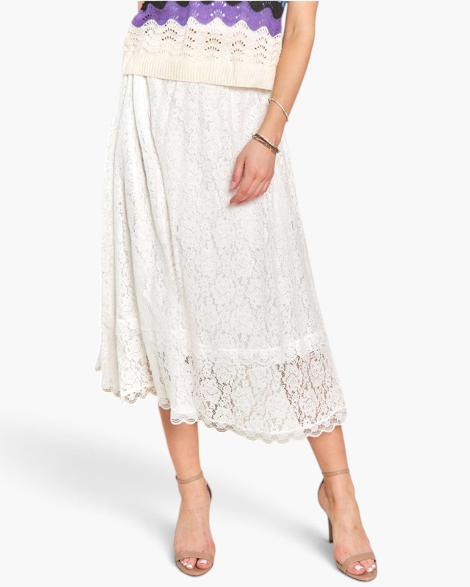 A model wearing a white lace midi skirt with a scalloped hem, paired with beige high-heeled sandals and a purple knit top, shown against a light gray studio background.