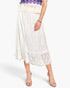 A model wearing a white lace midi skirt with a scalloped hem, paired with beige high-heeled sandals and a purple knit top, shown against a light gray studio background.