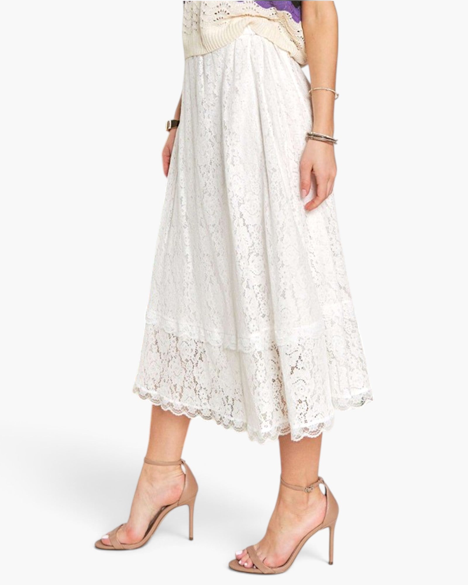 A model wearing a white lace midi skirt with a scalloped hem, paired with nude high-heeled sandals, shown from the waist down against a light gray studio background.