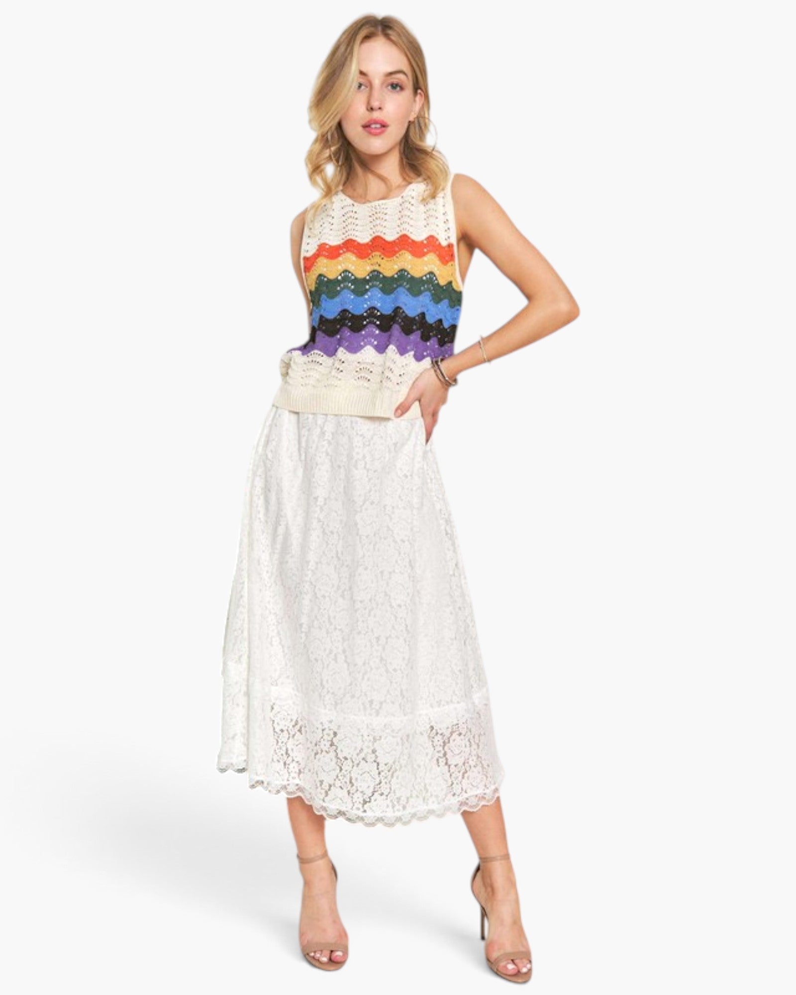 A woman modeling a sleeveless knit top with a rainbow stripe pattern and a white lace midi skirt, paired with nude high-heeled sandals against a light gray studio background.