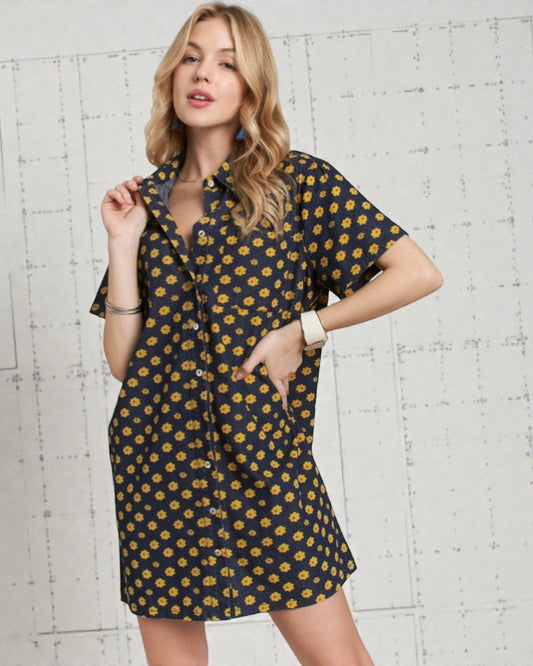 Woman wearing a navy dress with yellow floral pattern against a white brick wall.