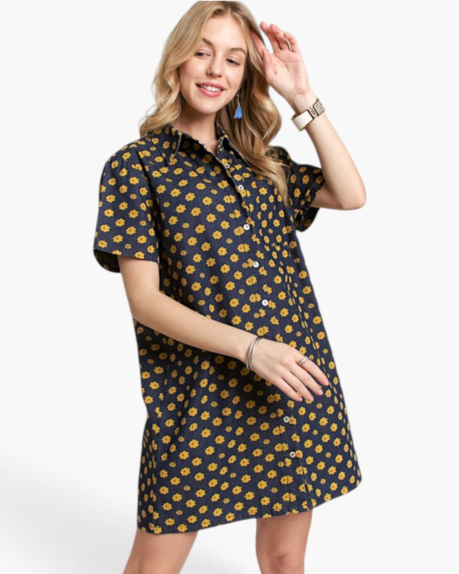 A woman modeling a short-sleeve button-up shirt dress with a dark navy blue background and a repeating yellow floral pattern, set against a plain light gray studio background.
