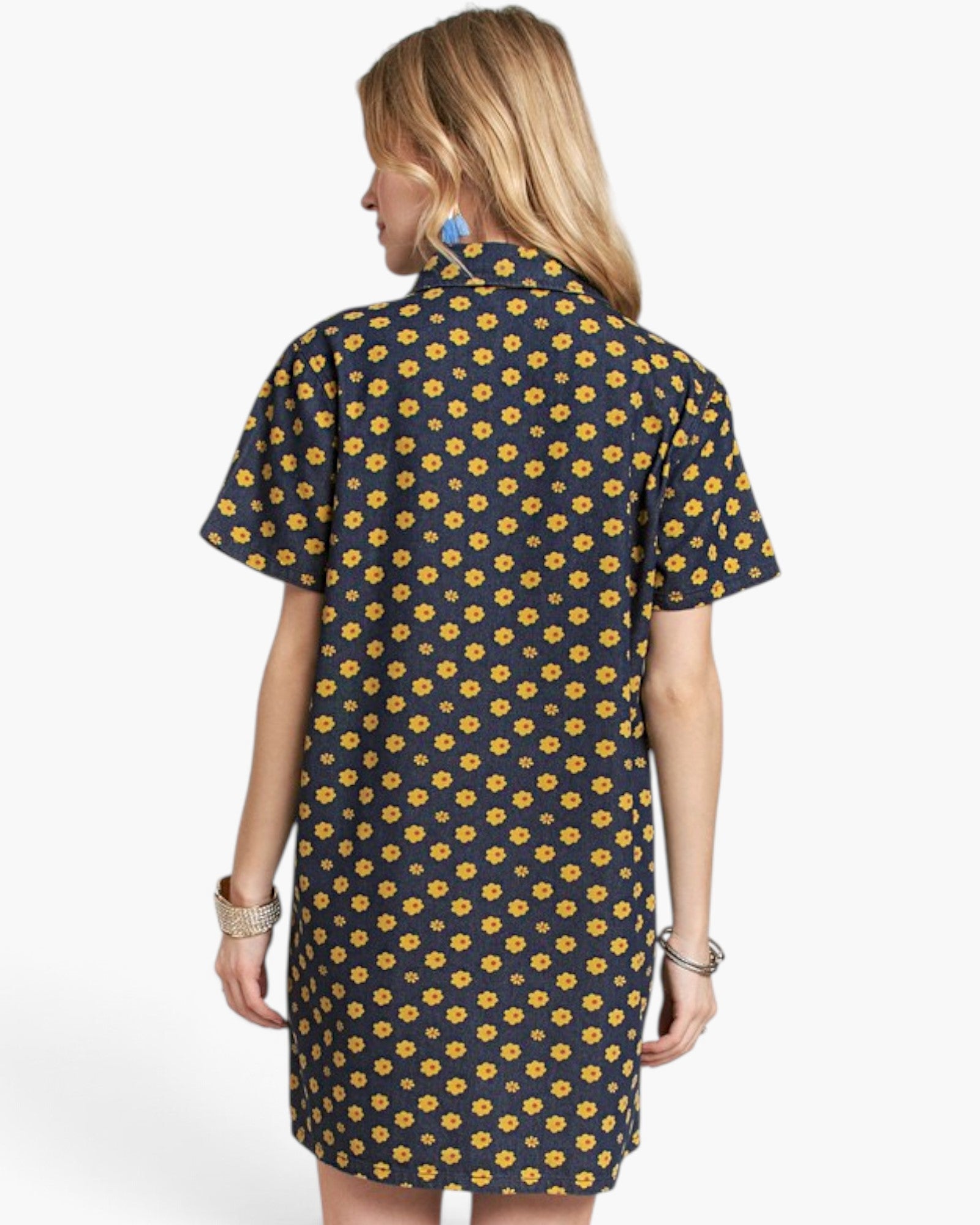 A woman shown from the back wearing a short-sleeve collared shirt dress with a repeating yellow flower print on a dark navy background, against a light gray studio background.