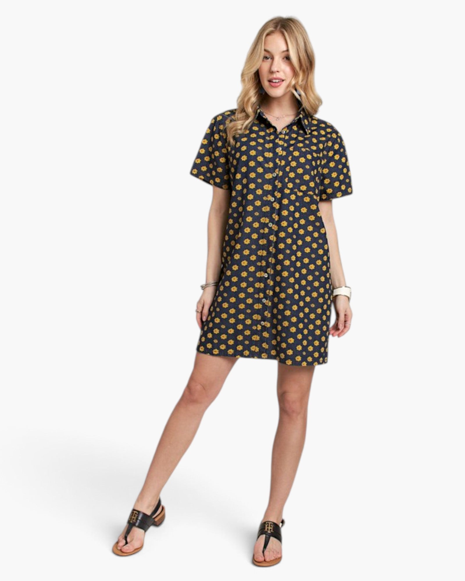 A woman modeling a short-sleeve button-up shirt dress with a navy blue base and a yellow floral pattern, paired with black thong sandals against a light gray studio background.