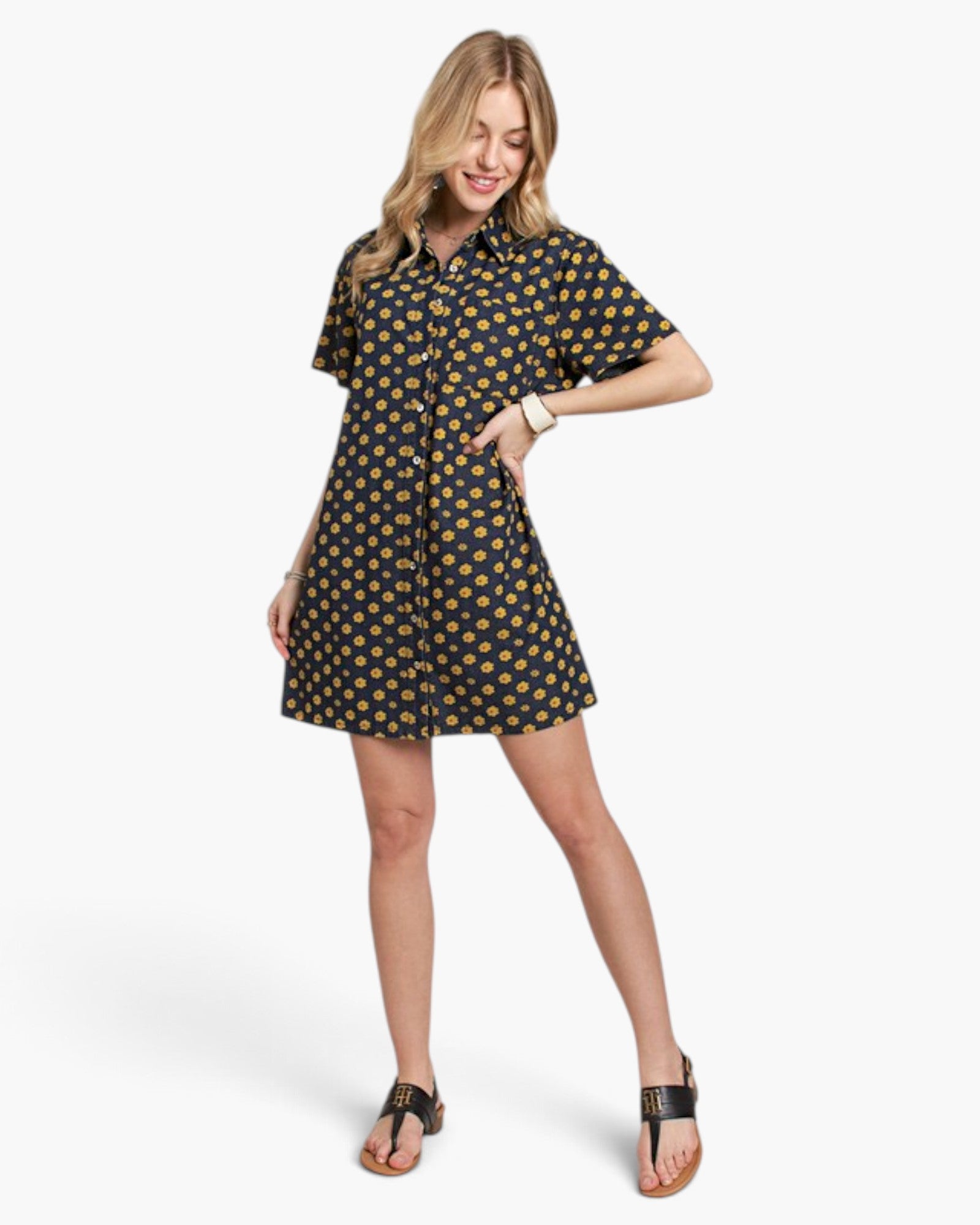A woman modeling a short-sleeve button-up shirt dress with a dark navy background and a repeating yellow and orange floral pattern, paired with black thong sandals against a light gray studio background.