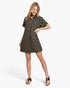 A woman modeling a short-sleeve button-up shirt dress with a dark navy background and a repeating yellow and orange floral pattern, paired with black thong sandals against a light gray studio background.