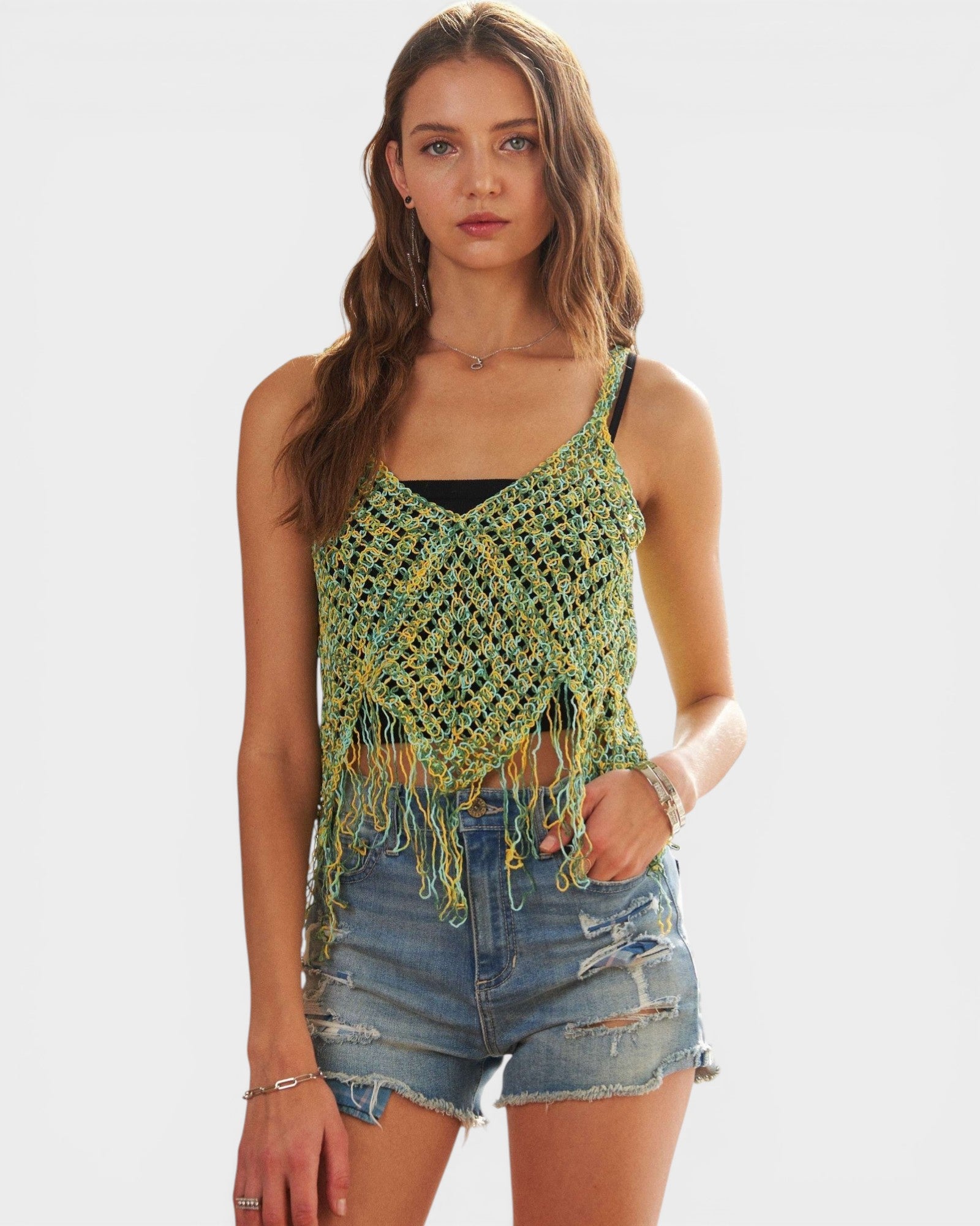 Woman wearing a green crochet tank top with fringe and denim shorts on a white background