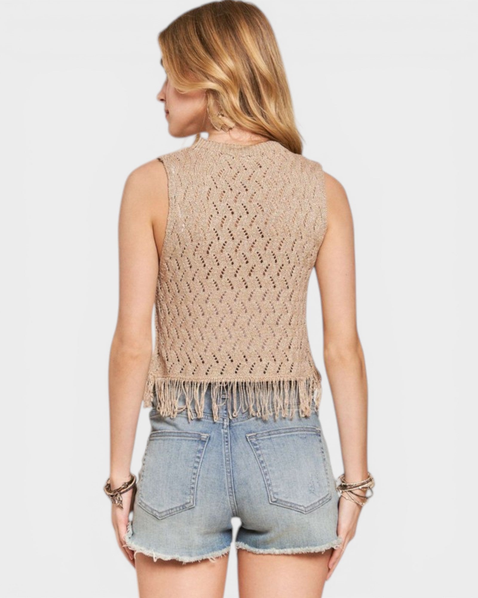 Woman wearing a beige sleeveless knitted top with fringe and denim shorts on a white background