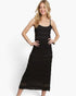A woman modeling a black sleeveless midi dress with thin spaghetti straps and a textured, ribbed knit body. The dress features a solid black bodice and a matching black lace hem, photographed against a clean white studio background.
