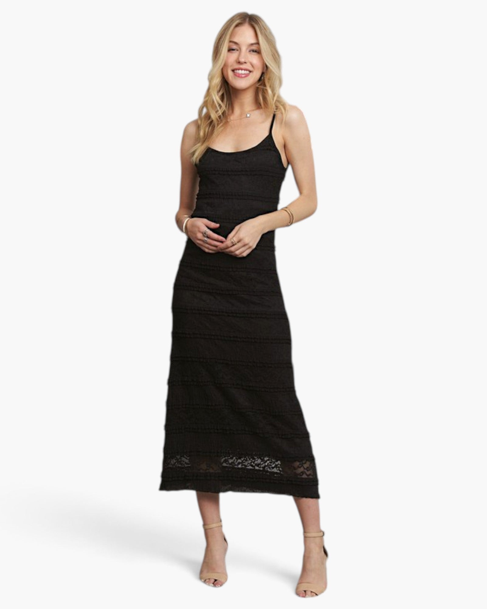 A woman modeling a black lace spaghetti-strap midi dress with a tiered skirt, paired with beige high-heeled sandals, against a plain white studio background.
