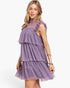 A woman modeling a lavender purple tiered tulle dress with ruffle details on the shoulders and a smocked bodice, set against a plain white background.