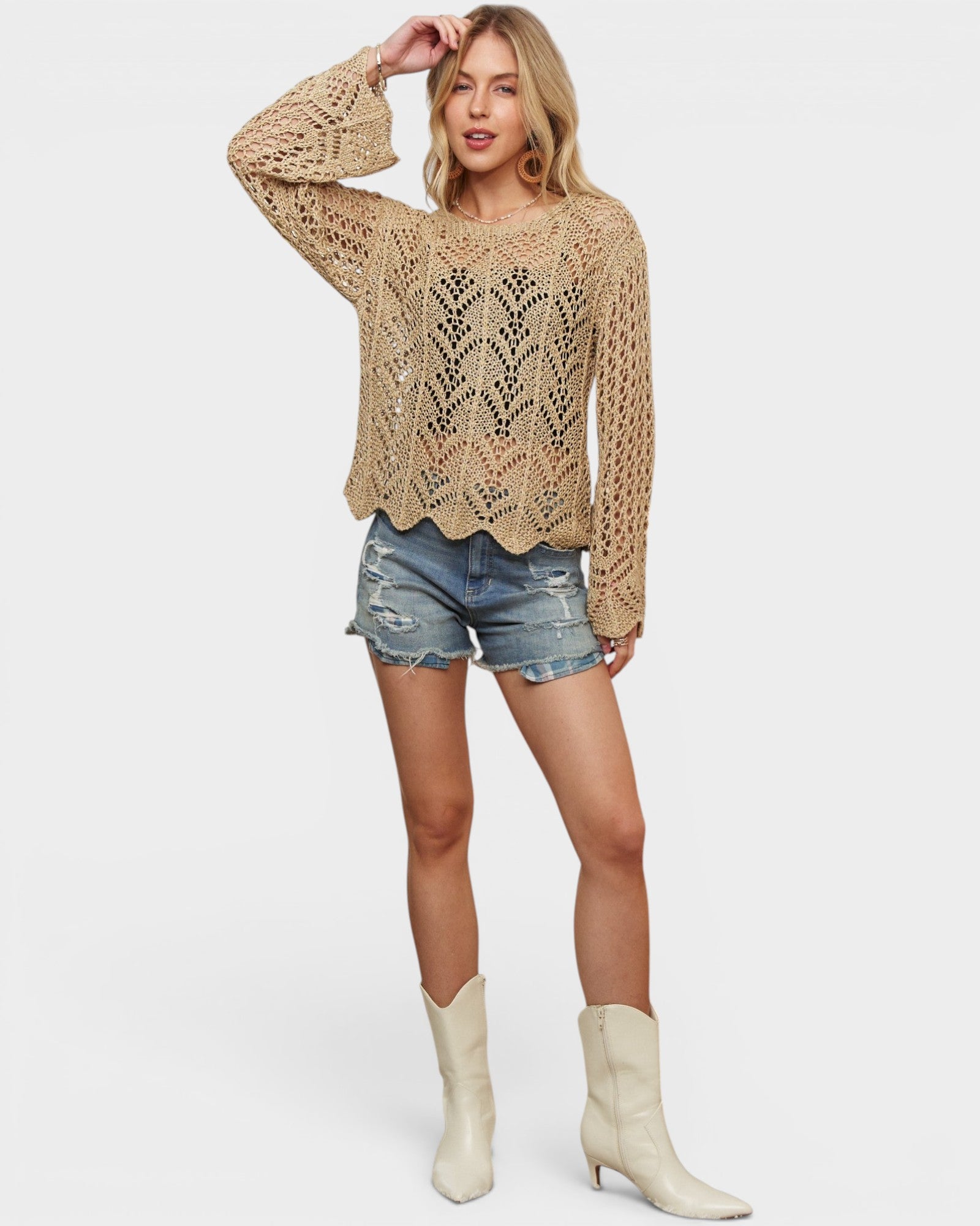 Woman wearing a beige crochet sweater, denim shorts, and white boots on a light background