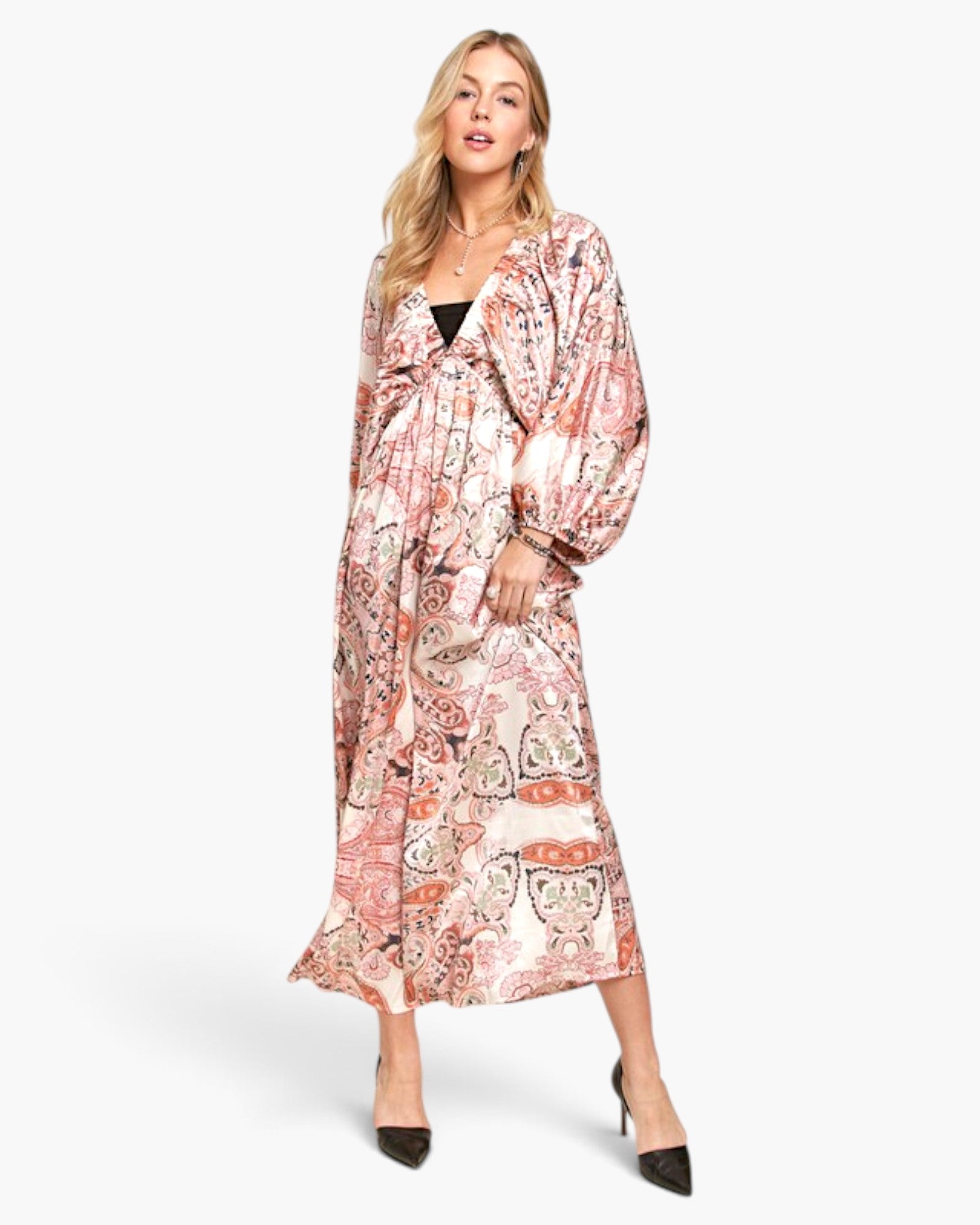 A woman modeling a long-sleeved midi dress with a pink, orange, and beige paisley print, styled with black pointed-toe heels against a light gray studio background.