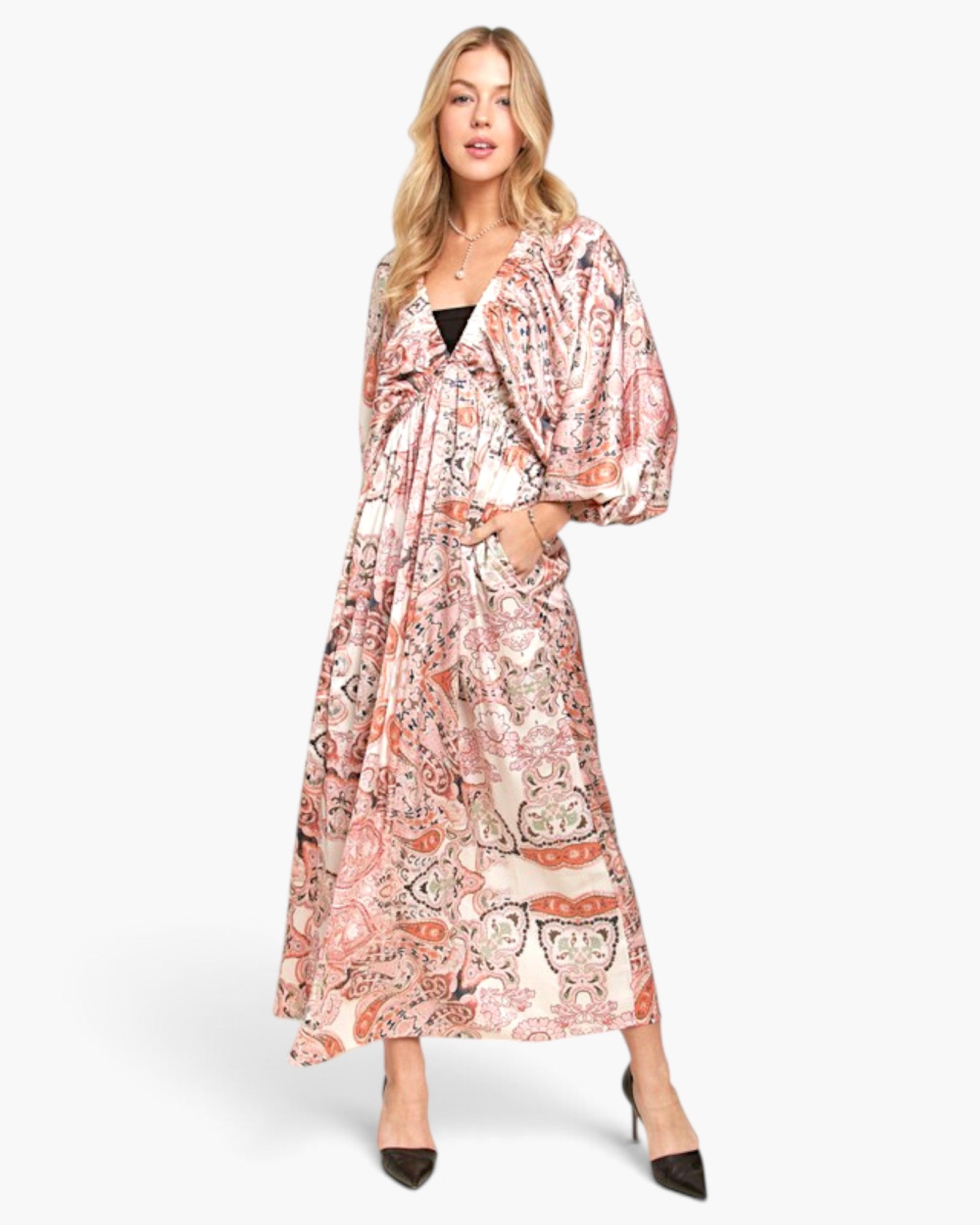 A woman modeling a long, flowing maxi dress with a pink, orange, and beige paisley pattern, styled with black pointed-toe heels against a plain white studio background.