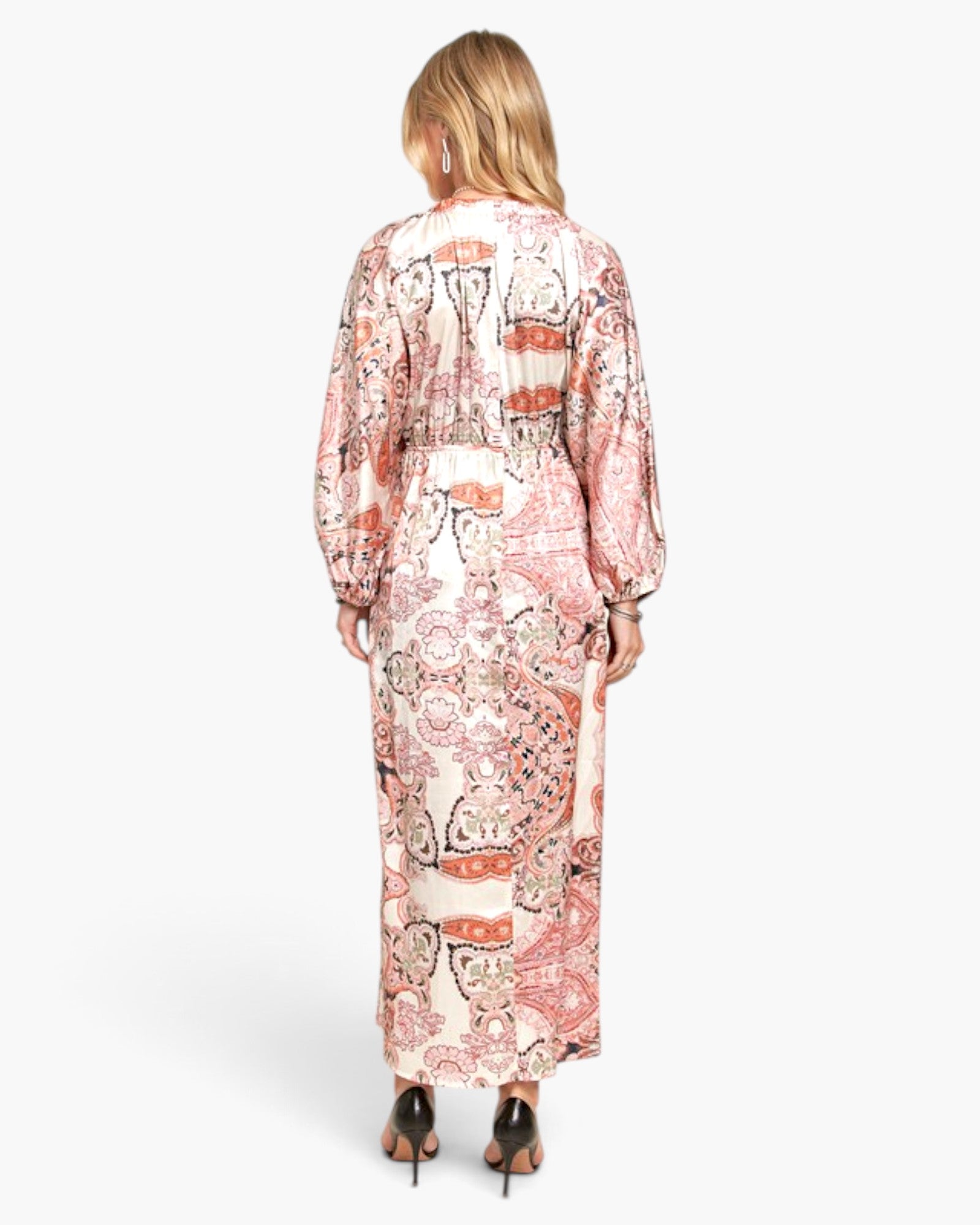 A woman viewed from the back wearing a long-sleeve maxi dress with a pink, orange, and black paisley pattern on a cream background, paired with black high heels, against a light gray studio background.