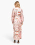A woman viewed from the back wearing a long-sleeve maxi dress with a pink, orange, and black paisley pattern on a cream background, paired with black high heels, against a light gray studio background.