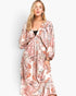A woman modeling a long-sleeve maxi dress with a paisley pattern in pink, orange, and beige tones over a black underlayer, set against a plain light gray background.