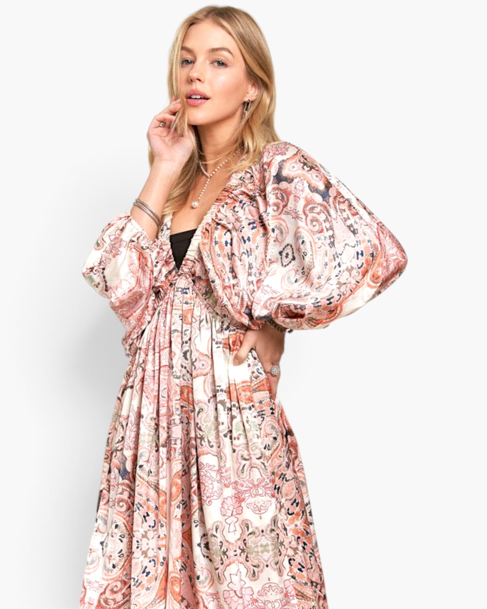 A woman modeling a long, loose bohemian maxi dress with voluminous balloon sleeves and an intricate paisley print in pink, beige, and black, set against a plain light gray background.