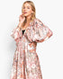 A woman modeling a long, loose bohemian maxi dress with voluminous balloon sleeves and an intricate paisley print in pink, beige, and black, set against a plain light gray background.