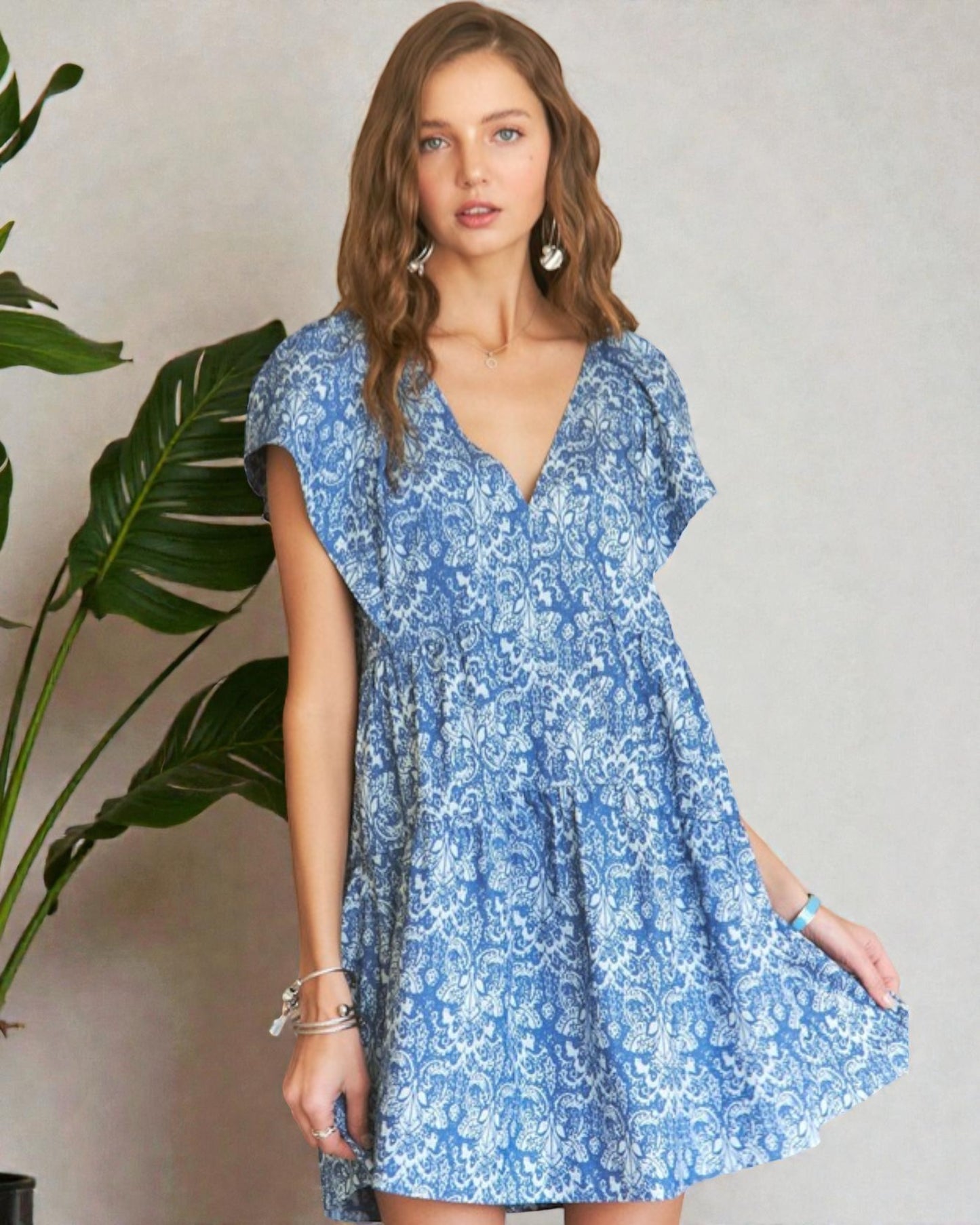 Woman wearing a blue floral dress standing next to a plant against a plain background
