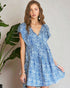 Woman wearing a blue floral dress standing next to a plant against a plain background