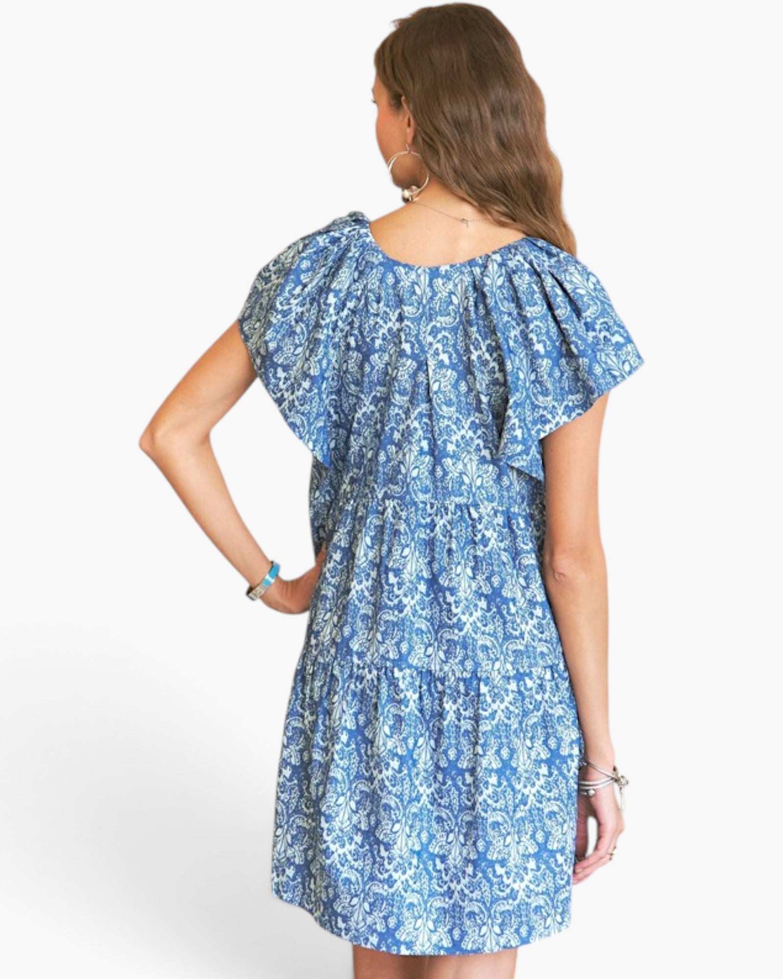 A woman shown from the back wearing a tiered blue and white patterned dress with flutter sleeves, photographed against a plain white studio background.