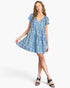 A woman modeling a short blue and white floral print dress with flutter sleeves and a V-neckline, paired with black heeled sandals against a light gray studio background.
