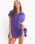 A woman modeling a purple short-sleeve button-up shirt with a textured crinkle fabric, paired with denim shorts, against a plain white background.