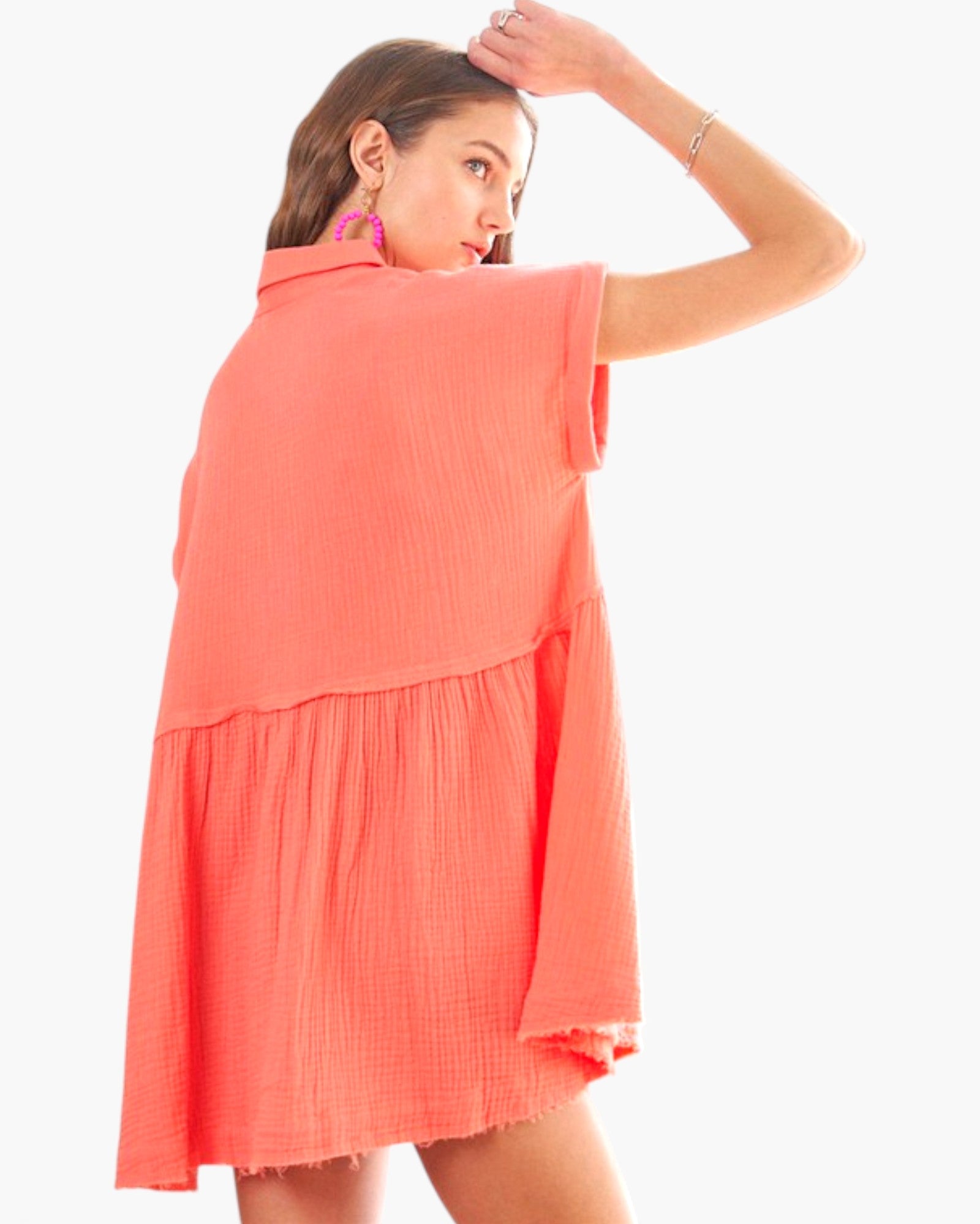 A woman modeling a short-sleeve coral pink textured dress with a tiered ruffle design, shown from a three-quarter back angle against a plain white background.